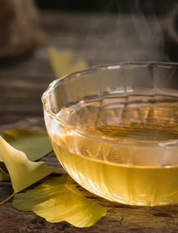 Chá de ginkgo biloba em xícara clara sobre mesa rústica, com folhas secas ao lado