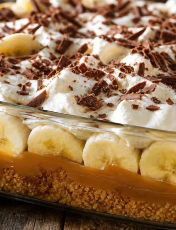 Torta banoffee na travessa com doce de leite, bananas, chantilly e chocolate sobre mesa de madeira